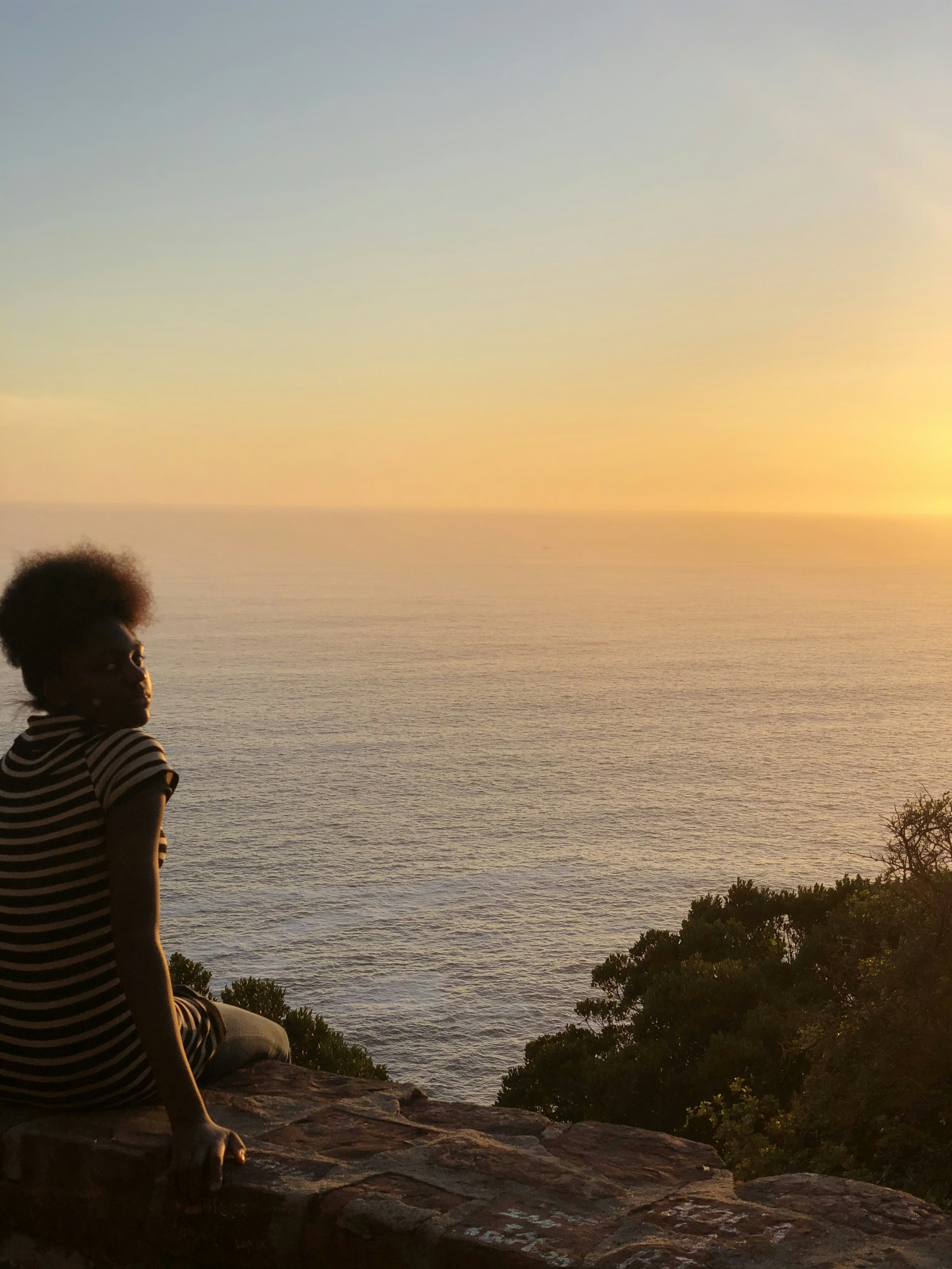 흑백 줄무늬 셔츠를 입은 여자는 일몰 동안 물가 근처의 바위에 앉아 있다