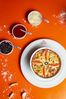 A small round pizza with slices of tomato, green peppers, and an olive on top is placed on a white plate. Surrounding the plate are small bowls containing different sauces, with blue-handled spoons resting in each. The background is a bright orange surface, sprinkled with some flour and a few cloves of garlic.