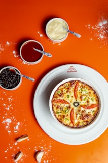 A small round pizza with slices of tomato, green peppers, and an olive on top is placed on a white plate. Surrounding the plate are small bowls containing different sauces, with blue-handled spoons resting in each. The background is a bright orange surface, sprinkled with some flour and a few cloves of garlic.
