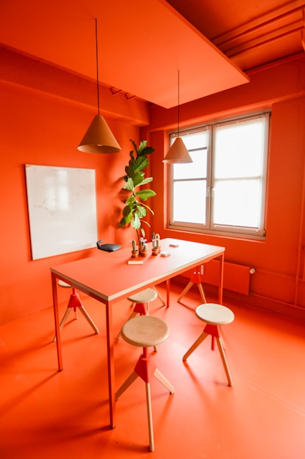A modern, minimalist room featuring orange walls and ceiling with matching orange furniture. A rectangular table is surrounded by three wooden stools with orange accents. Two cone-shaped pendant lights hang above the table. A whiteboard is mounted on the wall, and a tall plant is placed near the window, which lets in natural light.