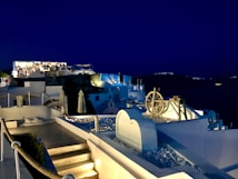 A nighttime view of a serene coastal village with whitewashed buildings and soft lighting illuminating terraces and pathways. The sky is a deep blue, and the architecture features classic Greek island elements, including arched roofs and a large decorative wheel.