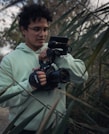 A person wearing a light green hoodie is holding a professional camera amidst tall green reeds. The individual is focused on their task, with the camera equipped with a grip and mounted with a small monitor. The setting appears to be outdoors with natural lighting.