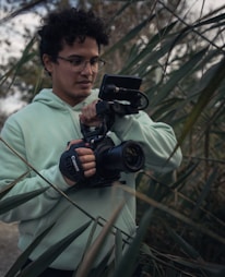 A person wearing a light green hoodie is holding a professional camera amidst tall green reeds. The individual is focused on their task, with the camera equipped with a grip and mounted with a small monitor. The setting appears to be outdoors with natural lighting.