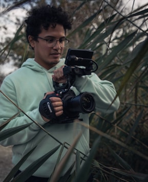 A person wearing a light green hoodie is holding a professional camera amidst tall green reeds. The individual is focused on their task, with the camera equipped with a grip and mounted with a small monitor. The setting appears to be outdoors with natural lighting.