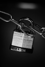 A close-up view of a metallic padlock secured to a chain. The padlock has horizontal engraved lines and the letters 'LSDA' on the base. The background is dark, highlighting the shiny, reflective surface of the lock and chain.