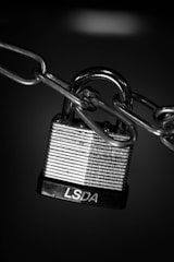 A close-up view of a metallic padlock secured to a chain. The padlock has horizontal engraved lines and the letters 'LSDA' on the base. The background is dark, highlighting the shiny, reflective surface of the lock and chain.