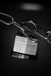 A close-up view of a metallic padlock secured to a chain. The padlock has horizontal engraved lines and the letters 'LSDA' on the base. The background is dark, highlighting the shiny, reflective surface of the lock and chain.
