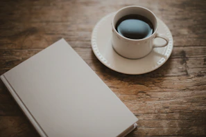 A rustic wooden table with a cup of coffee and a novel beside it.