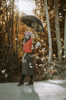 A woman stands on a pathway surrounded by autumn foliage. She is holding an open black umbrella and wearing a red top with a plaid skirt, black tights, boots, and a knitted hat. Fallen leaves are swirling around her as sunlight filters through the trees in the background, creating a warm glow.