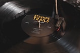 A close-up image of a black vinyl record being played on a turntable. The record label reads 'Strange Trails' by Lord Huron, with song titles listed. The turntable needle is positioned on the outer edge of the record, and there is a soft reflection on the surface.