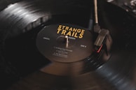 A close-up image of a black vinyl record being played on a turntable. The record label reads 'Strange Trails' by Lord Huron, with song titles listed. The turntable needle is positioned on the outer edge of the record, and there is a soft reflection on the surface.