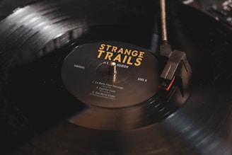 A close-up image of a black vinyl record being played on a turntable. The record label reads 'Strange Trails' by Lord Huron, with song titles listed. The turntable needle is positioned on the outer edge of the record, and there is a soft reflection on the surface.