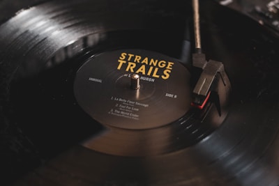 A close-up image of a black vinyl record being played on a turntable. The record label reads 'Strange Trails' by Lord Huron, with song titles listed. The turntable needle is positioned on the outer edge of the record, and there is a soft reflection on the surface.
