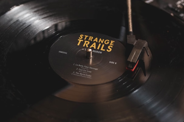 A close-up image of a black vinyl record being played on a turntable. The record label reads 'Strange Trails' by Lord Huron, with song titles listed. The turntable needle is positioned on the outer edge of the record, and there is a soft reflection on the surface.