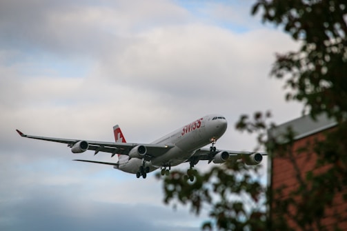 A commercial airplane with the logo and branding of a Swiss airline is flying low, preparing for landing. The sky is overcast with an array of clouds, and there are silhouettes of tree branches in the foreground.