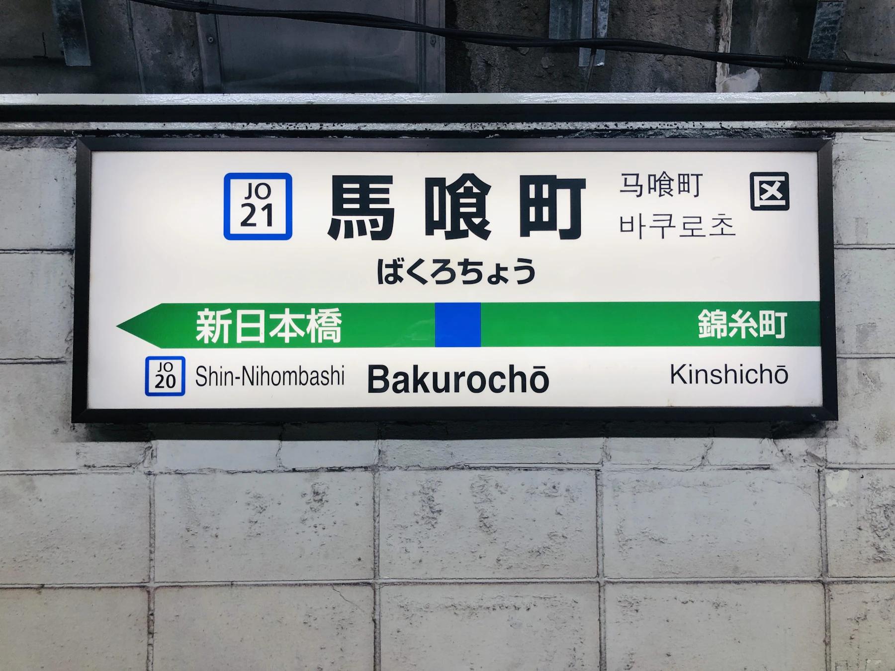 A Japanese railway station name board on a tiled wall, the station name in kanji, hiragana and Latin letters above a green band naming the next stops