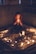 A person with long, wavy hair sits cross-legged in a dimly lit room, holding a candle in both hands. The setting features a tapestry on the floor, surrounded by an intricate circle of various objects and numerous lit candles, casting a warm glow.
