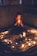 A person with long, wavy hair sits cross-legged in a dimly lit room, holding a candle in both hands. The setting features a tapestry on the floor, surrounded by an intricate circle of various objects and numerous lit candles, casting a warm glow.