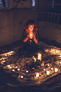 A person with long, wavy hair sits cross-legged in a dimly lit room, holding a candle in both hands. The setting features a tapestry on the floor, surrounded by an intricate circle of various objects and numerous lit candles, casting a warm glow.
