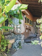 An outdoor area with lush green banana plants next to a rustic building made of stone and wood. The building features a unique, artistic design on the walls, including sculptural elements and earthy textures. There's a wooden storage box and a garden hose reel near the stone wall, and the ground is paved with gray bricks.