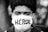 A close-up black and white photograph of a person wearing a white face mask with the word 'HÉROE' written on it. The background is blurred, highlighting the subject's face prominently.