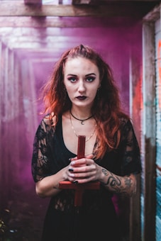 A person with red hair and gothic makeup stands in a dimly lit, narrow hallway with purple mist in the background. They are wearing a black lace outfit and have multiple tattoos on their arms. The person is holding a red cross in both hands.