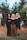 Three individuals stand in front of a backdrop of large evergreen trees. They are dressed in dark, gothic-style clothing with unique accessories and makeup, featuring dramatic eye designs. The setting is an overgrown area with grass and weeds, giving a sense of wilderness.