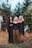 Three individuals stand in front of a backdrop of large evergreen trees. They are dressed in dark, gothic-style clothing with unique accessories and makeup, featuring dramatic eye designs. The setting is an overgrown area with grass and weeds, giving a sense of wilderness.
