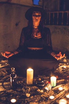 A person dressed in dark clothing and a wide-brimmed hat sits cross-legged on the ground, surrounded by candles and small decorative objects. The setting is dimly lit, creating a mysterious and mystical atmosphere.