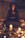 A person dressed in dark clothing and a wide-brimmed hat sits cross-legged on the ground, surrounded by candles and small decorative objects. The setting is dimly lit, creating a mysterious and mystical atmosphere.