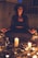 A person dressed in dark clothing and a wide-brimmed hat sits cross-legged on the ground, surrounded by candles and small decorative objects. The setting is dimly lit, creating a mysterious and mystical atmosphere.