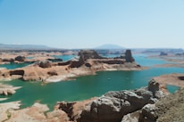 A vast lake with turquoise water is surrounded by rugged red rock formations under a clear blue sky. The rocky landscape consists of mesas and buttes, characteristic of a desert environment. The water gently curves around the rocky outcrops, creating a natural blend of land and water.