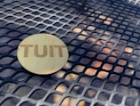 A wooden circular token with the word 'TUIT' engraved on it lies on a metal grid table with a geometric pattern.