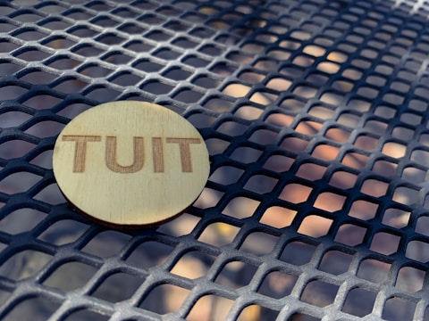 A wooden circular token with the word 'TUIT' engraved on it lies on a metal grid table with a geometric pattern.