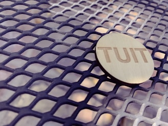 A round wooden token with the word 'TUIT' engraved on it rests on a metal mesh surface. The mesh creates a pattern of intersecting diamond shapes, and the lighting casts soft shadows.