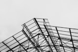 An intricate steel structure is prominently featured, with crisscrossing metal beams supporting transparent panels. The geometric pattern of the frame creates an industrial and modern aesthetic against a plain, overcast sky.