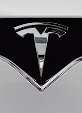 A close-up of a shiny metallic logo shaped like a stylized letter 'T' on a glossy black surface, bordered by light gray metal.