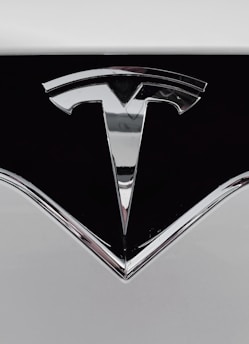 A close-up of a shiny metallic logo shaped like a stylized letter 'T' on a glossy black surface, bordered by light gray metal.