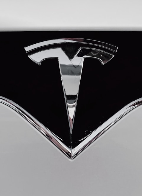 A close-up of a shiny metallic logo shaped like a stylized letter 'T' on a glossy black surface, bordered by light gray metal.