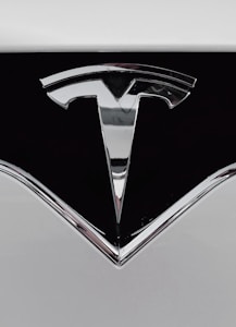 A close-up of a shiny metallic logo shaped like a stylized letter 'T' on a glossy black surface, bordered by light gray metal.