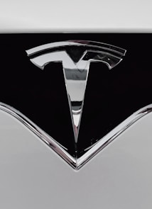 A close-up of a shiny metallic logo shaped like a stylized letter 'T' on a glossy black surface, bordered by light gray metal.