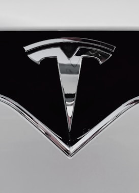 A close-up of a shiny metallic logo shaped like a stylized letter 'T' on a glossy black surface, bordered by light gray metal.