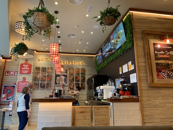 The image depicts the interior of a fast-casual restaurant with a rustic design. Wooden walls and ceiling feature prominently, with several hanging planters and decorative light fixtures. A counter with a menu board above features food images. A barista prepares drinks as two customers stand nearby, examining the menu.