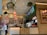 The image depicts the interior of a fast-casual restaurant with a rustic design. Wooden walls and ceiling feature prominently, with several hanging planters and decorative light fixtures. A counter with a menu board above features food images. A barista prepares drinks as two customers stand nearby, examining the menu.