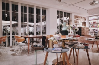 A cozy and modern interior of a cafe or bookstore, adorned with various objects like books, sculptures, and flowers. The setting includes several wooden tables and chairs, with large windows providing natural light. Artworks are displayed on the walls, adding a creative atmosphere.