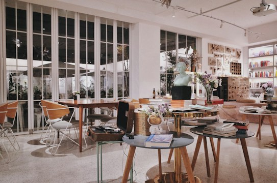 A cozy and modern interior of a cafe or bookstore, adorned with various objects like books, sculptures, and flowers. The setting includes several wooden tables and chairs, with large windows providing natural light. Artworks are displayed on the walls, adding a creative atmosphere.