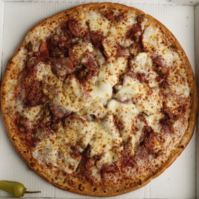 A large pizza covered with melted cheese and various meats, such as pepperoni and sausage, placed on a white cardboard surface. A single whole pepper is positioned near the bottom left corner.