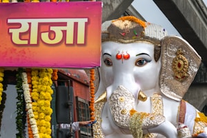 A large, ornately decorated statue of the Hindu deity Ganesha is positioned in the center. The statue is adorned with floral garlands, particularly marigolds, and has detailed embellishments on its trunk, ears, and head. To the left, there is a bright pink and orange sign with fancy script. The background reveals parts of a building structure and what appears to be an elevated walkway or bridge.