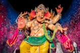 A colorful statue of a multi-armed deity adorned with jewelry and elaborate clothing, set against a vibrant, starry background. The deity has the face of an elephant and a small symbol on its palm, with various accessories enhancing its divine appearance.