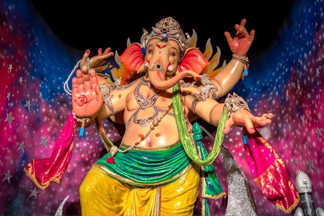 A colorful statue of a multi-armed deity adorned with jewelry and elaborate clothing, set against a vibrant, starry background. The deity has the face of an elephant and a small symbol on its palm, with various accessories enhancing its divine appearance.